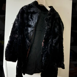 Svart pälsjacka i fuskpäls - Mysig svart pälsjacka i fuskpäls med klassisk krage och helsvart foder. Jackan har en rak passform och är perfekt för dig som vill ha en stilren och trendig look under kyliga dagar.