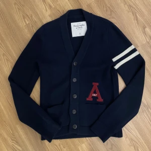 Mörkblå kofta Abercrombie & Fitch  - Mörkblå kofta från Abercrombie & Fitch i storlek S med knappar framtill, två fickor och röd A-logga på bröstet. Ärmen har två vita ränder som cool detalj. Koftan är stickad och har en klassisk, lite preppy stil som passar perfekt till jeans. (Modell 176cm, 70kg)
