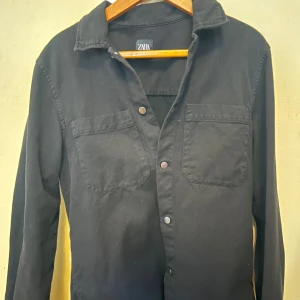 Svart overshirt från Zara - Svart overshirt från Zara i bomull med klassisk krage, två bröstfickor och knappar framtill. Perfekt att slänga över en t-shirt för en chill look. Långärmad och enkel design som funkar till det mesta.