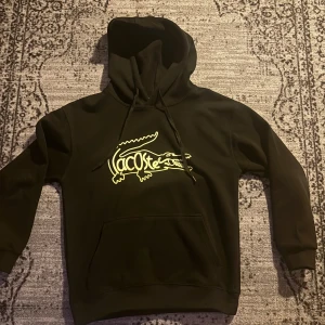 Svart hoodie från Lacoste med logga - Svart hoodie från Lacoste med stor gul krokodil-logga på bröstet. Klassisk huva med snörning och magficka framtill. Perfekt för dig som gillar streetwear och vill sticka ut med en ikonisk look. Pris kan diskuteras 