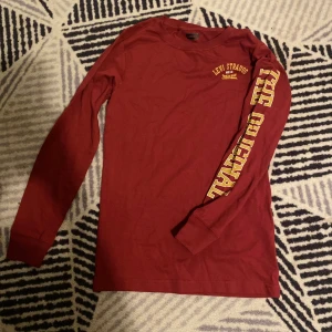 Vinröd långärmad Levi's tröja - Snygg vinröd långärmad tröja från Levi's med gul texttryck på bröstet och längs ena ärmen. Klassisk crewneck och skön passform i mjuk bomull. Perfekt för dig som gillar streetstyle och vill ha något bekvämt men ändå stilrent.