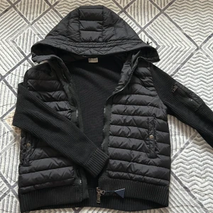 Svart pufferjacka från Moncler - Svart pufferjacka från Moncler med stickade ärmar och quiltad kropp. Jackan har huva, dragkedja framtill och Moncler-logga på ärmen. Snygga detaljer som ribbade muddar och fickor med dragkedja och knapp. Perfekt för dig som vill ha en stilren och sportig look.