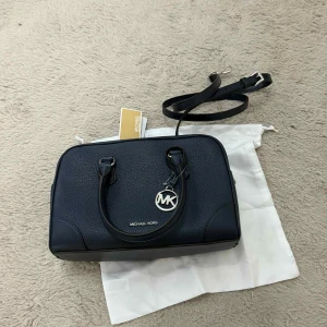 Michael Kors Handväska - Helt Ny - Elegant och kompakt handväska från Michael Kors i mörkblått med klassisk MK-detalj framtill. Väskan är helt ny och oanvänd.  Detaljer: • Färg: Mörkblå • Material: Slitstarkt konstläder • Justerbar axelrem + handtag • Flera fack inuti för nycklar, mobil och mindre tillbehör • Stilren och smidig design som passar både vardag och fest  En lyxig, mindre väska i hög kvalitet – perfekt för dig som vill ha något elegant och praktiskt.