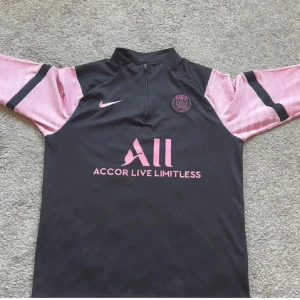 Svart och rosa PSG fotbollströja Nike - Snygg PSG fotbollströja från Nike med svart kropp och ljusrosa långärmade ärmar med svarta detaljer. Tröjan har halv dragkedja, PSG-logga och sponsortryck i rosa framtill. Perfekt för dig som gillar sportig stil och Paris Saint-Germain.