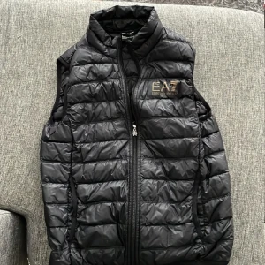 Svart dunväst från Emporio Armani EA7 - Säljer en svart dunväst från Emporio Armani EA7 med quiltad design och dragkedja framtill. Västen har hög krage och logotyp på bröstet. Perfekt för lager-på-lager och har en clean, sportig look.
