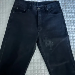 Svarta jeans från Chrome Hearts - Svarta jeans från Chrome Hearts med ikoniska korsapplikationer i läder på både fram- och baksida. Snygga silverfärgade knappar och detaljer, klassisk femficksmodell och raka ben. Jeansen är tillverkade i kraftig denim och har en unik streetstil.
