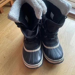 Svarta vinterstövlar från Sorel - Svarta vinterstövlar från Sorel med vit fuskpälsfoder, vattentätt material och grov sula för bra grepp. Klassisk design med snörning och logga på sidan. Perfekta för kalla dagar och snö, håller fötterna varma och torra.