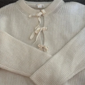 Beige stickad kofta med knyt - Superfin beige stickad kofta från Wheat med tre dekorativa knytband framtill. Mjuk och mysig, perfekt för lager-på-lager under kyliga dagar. Rund halsringning och långa ärmar ger en klassisk look.