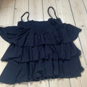 Svart volangtopp från Second Female - Supersnygg svart topp från Second Female med flera lager volanger och smala axelband. Modellen är luftig och har en flowy känsla, perfekt för dig som gillar en trendig och feminin stil. Toppen är tillverkad i mjuk bomull och passar perfekt till jeans eller kjol.