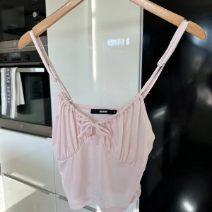 Ljusrosa linnetopp från BikBok🌸 - Supersöt ljusrosa linnetopp från BikBok med smala axelband och en liten rosett framtill. Toppen har en lätt rynkad detalj vid bysten som gör att man kan justera hur tajt man vill knyta🩷