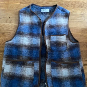 Rutig ullväst från Universal Works - Riktigt snygg rutig väst i blått, brunt och grått från Universal Works. Västen är gjord i ull, har bruna kantband, tre fickor och stängs med dragkedja. Perfekt lager-på-lager-plagg med cool vintagekänsla.