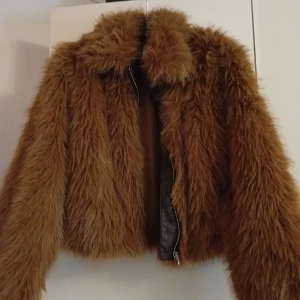 Brun pälsjacka med dragkedja - Säljer en brun pälsjacka med fluffig, långhårig fuskpäls och markerad krage. Jackan är croppad och har dragkedja framtill. Perfekt för dig som vill sticka ut och hålla dig varm med stil.
