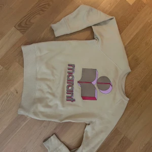 Beige sweatshirt från Isabel Marant - Säljer en beige sweatshirt från Isabel Marant Étoile med ikoniskt tryck i rosa, rött och grått framtill. Tröjan har rund hals, långa ärmar och är gjord i en mjuk bomullsblandning med polyester. Perfekt för en chill och trendig look.