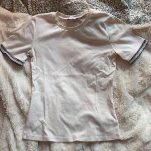 Vit topp med glittriga ärmslut från Zara - Snygg vit topp från Zara med korta ärmar och rund hals. Ärmsluten har en glittrig silverdetalj och svart kant som ger en extra touch. Toppens passform är normal och materialet känns mjukt och bekvämt, perfekt till jeans eller kjol.