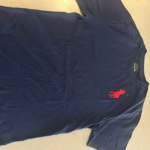 Mörkblå t-shirt Polo Ralph Lauren XL - Snygg mörkblå t-shirt från Polo Ralph Lauren i storlek XL. T-shirten har ett stort rött broderat polospelar-motiv på bröstet och en röd broderad detalj på ärmen. Tillverkad i mjuk bomull och har klassisk passform.