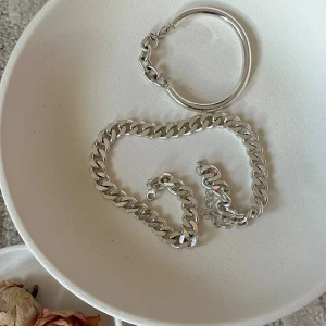 Set med silverfärgade kedjesmycken - Säljer ett set med chunky silverfärgade smycken: ett armband med kraftig kedja, ett matchande halsband och ett stilrent stelt armband. Perfekt för dig som gillar statement-accessoarer och vill sticka ut med en clean look.