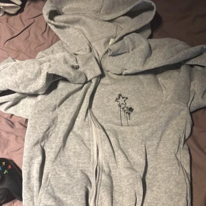 Grå hoodie med dragkedja och stjärntryck - Grå hoodie utan märke med hel dragkedja och huva. På bröstet finns ett svart tryck med stjärnor. Tröjan har långa ärmar och är tillverkad i mjukt bomullsmaterial. Helt oanvänd. 