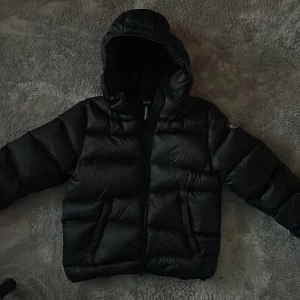 Svart pufferjacka från Colmar - Säljer min sons svarta pufferjacka från Colmar med huva och dragkedja. Jackan är vadderad och har en klassisk Colmar-logga på ärmen. Perfekt för kalla dagar och riktigt snygg till streetwear-stilen.