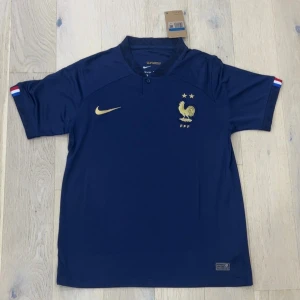 Frankrike Nike fotbollströja marinblå - Marinblå officiell Frankrike fotbollströja från Nike med guldiga detaljer, FFF-logga och tupp på bröstet. Kortärmad modell i lätt och ventilerande material. Små franska flaggor på ärmsluten ger extra stilpoäng.