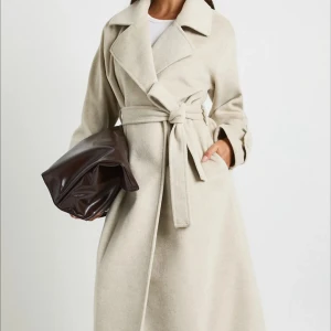 Beige kappa med bälte och stora slag - Snygg beige kappa med breda slag, knytskärp i midjan och dekorativa knappar vid ärmsluten. Jackan har en rak och lång siluett, sidfickor och är tillverkad i ett mjukt ullmaterial. Perfekt för dig som vill ha en stilren och klassisk look. Den håller värmen och är bra inför höst/vinter. Är i storlek M/38 men passar XS-L. Ordinarie pris 1370:- kommer i sin egna förpackning samt prislapp