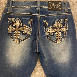 Blå jeans med broderade fickor - Snygga blå skinny jeans från Cello med broderade detaljer och nitar på bakfickorna. Jeansen har kontrastsömmar, slitningar vid fickorna och en tight passform. Perfekta för dig som gillar unika detaljer och vill sticka ut. Snyggt om man klipper till shorts. Midja rakt: 34cm Lår: 23cm Innerben: 81cm