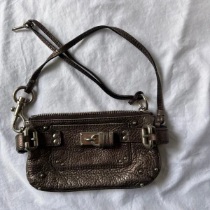 Chloé brun handväska i skinn - Cool brun axelväska från Chloé i äkta skinn med metallic finish. Väskan har snygga silverdetaljer, spännen och ett ikoniskt lås framtill. Praktisk dragkedja upptill och justerbar axelrem. Perfekt för dig som vill ha en statementväska med lyxig känsla.