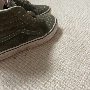 Olivgröna Vans Sk8-Hi sneakers - Säljer ett par olivgröna Vans Sk8-Hi sneakers med klassisk vit sula och vit sidestripe. Skorna har hög modell, snörning och är tillverkade i mocka med detaljer i textil. Perfekta för dig som gillar streetstyle och vill ha en ikonisk look.