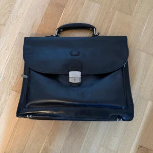 Baron Portfölj - Klassisk svart portfölj i skinn från Baron med stilren design. Väskan har ett robust handtag, silverfärgat lås och dragkedjefack på sidan. Perfekt för att bära laptop, papper och andra viktiga grejer till plugget eller jobbet. 10000 nypris! 