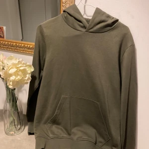 Olivgrön hoodie med ficka - Säljer en olivgrön hoodie med klassisk känguruficka framtill och huva. Tröjan har långa ärmar och ribbade muddar vid ärmslut och nederkant. Perfekt för chill dagar eller när du vill ha en enkel och stilren look. i använt fint skick