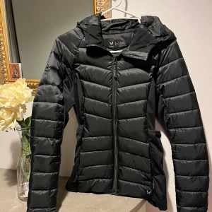 Svart pufferjacka från SOC med huva - Svart pufferjacka från SOC med snygg figurnära passform och huva. Jackan har dragkedja framtill, två fickor med dragkedja och quiltade paneler. Materialet är polyester och jackan är perfekt för kyliga dagar.