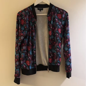 Blommig bomberjacka Way of Sweden - Snygg bomberjacka från Way of Sweden med färgstarkt blommönster i rött och blått på svart botten. Jackan har svarta muddar, dragkedja framtill och klassisk bomberkrage. Perfekt för dig som vill sticka ut med en cool och unik stil.