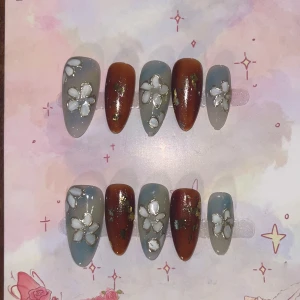 XS - Blommiga lösnaglar i blått och brunt - Snygga lösnaglar i mandelform med mix av blågrå och brun bas, dekorerade med vita blommor och guldiga detaljer. Perfekt för dig som vill ha en unik och kreativ nageldesign. Kommer i set om tio stycken.