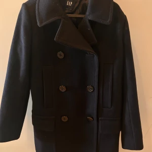 Svart skepparkavaj från GAP - Klassisk svart skepparkavaj från GAP i ullblandning. Jackan har dubbelknäppning med stora knappar, bred krage och raka fickor framtill. Storlek S men har lite större passform så skulle lätt passa dig som bär storlek 38\M