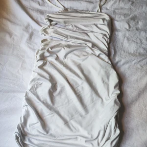 Vit omlott kortklänning med smala axelband - Säljer en vit kortklänning med smala axelband och snygga draperade detaljer längs sidorna. Klänningen har omlott-design och är tillverkad i ett mjukt, stretchigt material som sitter skönt mot kroppen. Perfekt för dig som gillar minimalistisk och stilren look.