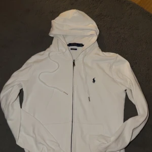 Vit hoodie från Polo Ralph Lauren - Vit hoodie från Polo Ralph Lauren med dragkedja och huva. Klassisk design med broderad logga på bröstet och snörning i huvan. Tillverkad i mjuk bomull och har en normal passform. Perfekt för en clean och stilren look.