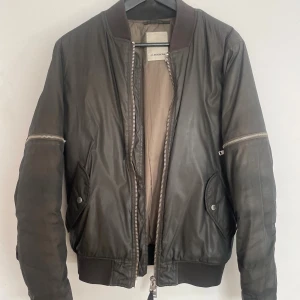 bomberjacka från J.Lindeberg - Snygg svart bomberjacka från J.Lindeberg med klassisk ribbad krage, muddar och midja. Jackan har dragkedjor både framtill och på ärmarna samt två fickor med tryckknappar. Materialet är slätt och ger en stilren look.