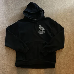 Svart hoodie från Hoodrich UK M - Svart hoodie från Hoodrich UK med stor vit tryckt text och grafiskt motiv på ryggen samt mindre tryck på bröstet. Klassisk känguruficka, huva och ribbade muddar. Perfekt streetwear-vibe och riktigt snygg passform.