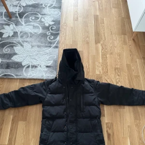 Svart dunjacka från Canada Goose - Säljer en svart dunjacka från Canada Goose med ikonisk patch på ärmen och stor huva med äkta päls.                                                    Det finns tydliga tecken på användning.                      Höger fickan saknar dragkedja och kroken på jackkedjan i mitten saknas.                                   Det går att fixa hos en skräddare.                         Priset kan diskuteras. 
