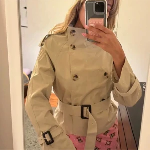 Beige trenchcoat med bälte och knappar - Snygg beige trenchcoat med dubbelknäppning, höga axlar och bälte i midjan. Jackan har klassiska detaljer som axelklaffar och spännen vid ärmsluten. Perfekt för dig som vill ha en stilren och tidlös look.