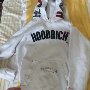 Vit hoodie från Hoodrich med tryck - Vit hoodie från Hoodrich med stor svart och röd logga på bröstet och detaljerat tryck på ryggen. Huva med tryck och känguruficka framtill. Mjuk och skön, perfekt för streetwear-stil.