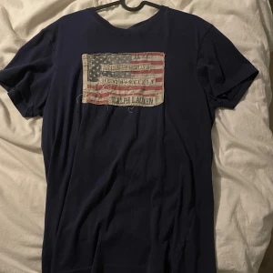 Mörkblå t-shirt Ralph Lauren S - Mörkblå t-shirt från Ralph Lauren Denim & Supply med en stor patch av amerikanska flaggan på bröstet. T-shirten har rund hals, korta ärmar och är gjord i mjuk bomull. Perfekt för dig som gillar en cool och avslappnad stil.