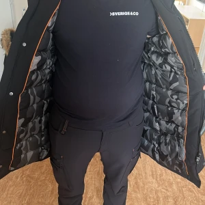 Svart jacka från Hollies med äkta päls  - Säljer en svart jacka från Hollies med snygg äkta päls  på huvan och camomönstrat innerfoder. Jackan har dragkedja, knappar och flera fickor. Perfekt för kalla dagar med sin långa modell och varma känsla. Stilsäker badge på ärmen. (PRIS GÅR ATT DISKUTARA) 