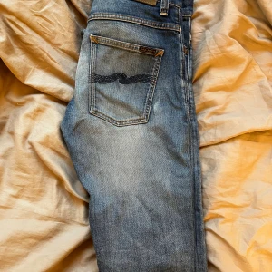 Blå jeans från Nudie Jeans, W32 L36 - Säljer ett par klassiska blå jeans från Nudie Jeans med snyggt slitna detaljer och kontrastsömmar. Modellen har fem fickor och Nudie-loggan på bakfickan. Perfekt passform och riktigt skön denimkänsla.