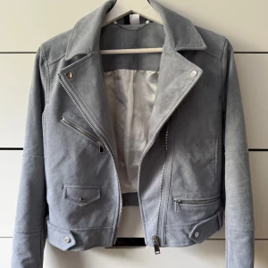 Mocka bikerjacka med dragkedjor - Snygg grå bikerjacka i mocka-look med silvriga dragkedjor och klassisk krage. Aldrig använd. Satin liknande material inuti. Liten I storlek.