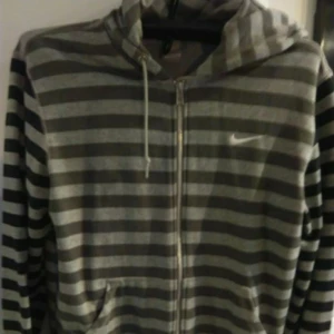 Randig hoodie från Nike med dragkedja - Grå och svart randig hoodie från Nike med dragkedja framtill, huva med snörning och två fickor. Perfekt för chill dagar eller när du vill ha en sportig look. Mjukt material och klassisk Nike-logga på bröstet.