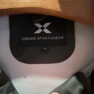 Svart regnjacka Cross Sportswear XS - Svart regnjacka från Cross Sportswear i storlek XS. Jackan har huva med vita snören, dragkedja och tryckknappar framtill samt två snedställda fickor. Tillverkad i syntetmaterial som står emot regn, perfekt för ruggiga dagar.