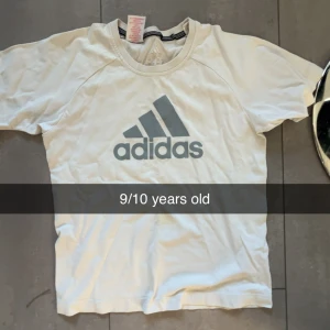 Vit Adidas t-shirt med logga - Vit t-shirt från Adidas med klassisk stor grå logga på bröstet. T-shirten har rund halsringning och korta ärmar. Perfekt för dig som gillar sportig och enkel stil.