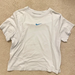  Nike t-shirt med blå logga - Ljusblå t-shirt från Nike med klassisk blå swoosh broderad på bröstet. Enkel och clean design med rund hals och korta ärmar. Perfekt för en sportig och avslappnad stil.