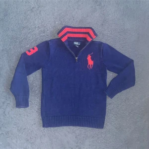 Blå half-zip från Ralph Lauren - Mörkblå stickad tröja från Ralph Lauren med röd broderad logga på bröstet och röd siffra 3 på ärmen. Tröjan har ribbade muddar, halv dragkedja och en krage med röda ränder på insidan. Perfekt för dig som gillar klassisk och sportig stil. OBS: den är i M för barn, vilket är runt xs så utgå från måtten