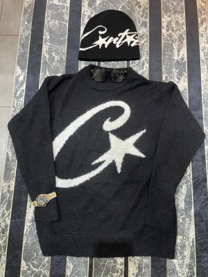 CORTIEZ & MÖSSA  - Svart stickad tröja från Corteiz med en stor vit grafisk stjärna och C på framsidan. Rund hals och långa ärmar, loose fit. Perfekt för dig som gillar streetwear och vill sticka ut med en fet design.